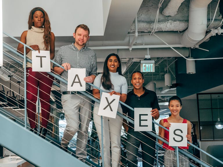 A diverse group of professionals smiling and holding 'TAXES' signs on an indoor stairway